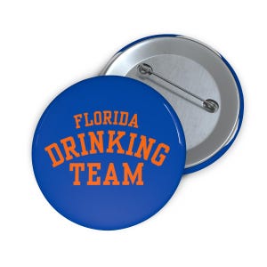 Peut inclure: Un bouton bleu avec les mots "FLORIDA DRINKING TEAM" en orange. Le bouton a une épingle en métal à l'arrière et un bord argenté brillant. Un accessoire amusant.