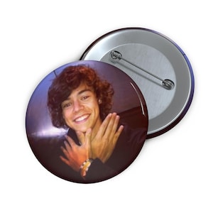 May include: A round button pin featuring a smiling person with curly brown hair, hands clasped in front. The pin has a silver metal back with a pin clasp. The image has a purple and orange color scheme.