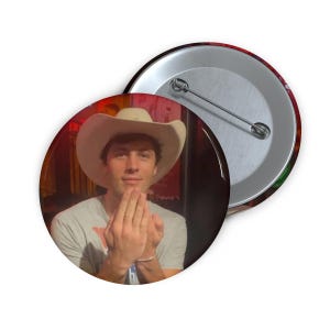 May include: A round button featuring a person wearing a cowboy hat. The button has a silver pin back. The person is wearing a gray t-shirt. The background is blurred with red and brown tones.