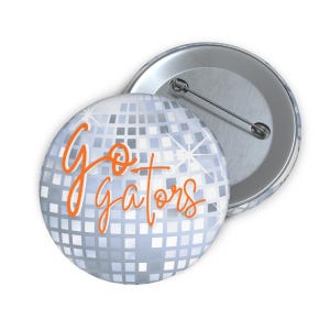 Peut inclure: Un bouton rond avec un motif de boule disco argentée et l'inscription "Go Gators" en orange. Le bouton est doté d'une épingle au dos et se détache sur un fond blanc. Article pour les fans.
