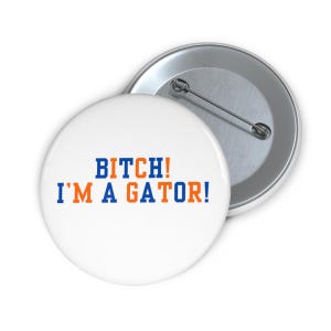 Peut inclure: Bouton blanc avec le texte "BITCH! I'M A GATOR!" en lettres bleues et oranges. Le bouton a un dos en métal et une épingle. Le design est une déclaration audacieuse.