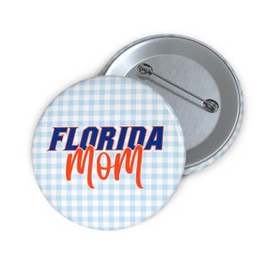 Peut inclure: Un bouton rond avec un motif vichy bleu clair et blanc. Le bouton porte l'inscription "FLORIDA" en bleu et "MOM" en orange. Un deuxième bouton est visible en arrière-plan.