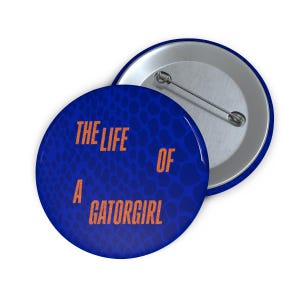 Peut inclure: Deux badges ronds. L'un est face visible, l'autre est face cachée. Le devant du badge est bleu avec un motif en forme d'écailles. Le texte "THE LIFE OF A GATORGIRL" est imprimé en orange. Le dos du badge est argenté.