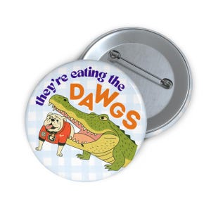Peut inclure: Un bouton rond avec une illustration de dessin animé d'un alligator mangeant un bouledogue portant un maillot rouge. Le texte "they're eating the DAWG" est imprimé sur le bouton. Le fond est un motif à carreaux bleu clair et blanc.