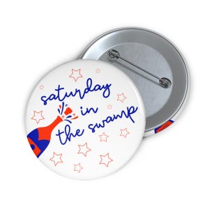 Peut inclure: Un badge blanc avec l'inscription "saturday in the swamp" en bleu, une illustration de bouteille de champagne rouge et bleu et des étoiles rouges. Un autre badge est également visible.