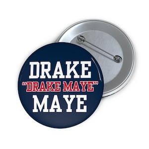May include: A round button with the text "DRAKE MAYE" in white and red lettering on a navy blue background. A second button is visible in the background, with a silver pin.