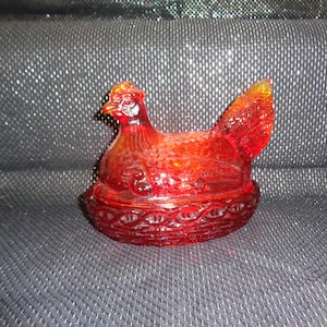 May include: A red glass hen-shaped dish with a basket weave base. The hen has a detailed comb and tail, and the glass has a textured, translucent appearance. This decorative piece is likely vintage.