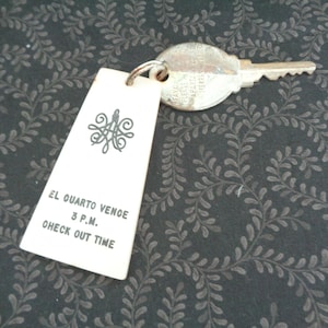 May include: A vintage hotel key. The metal key is attached to a cream-coloured tag with the text "EL QUARTO VENCE 3 P.M. CHECK OUT TIME" and a decorative emblem. The key and tag are on a dark background with a leaf pattern.