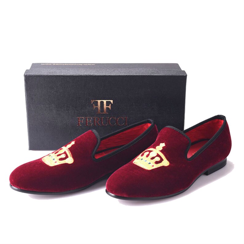 May include: A pair of burgundy velvet loafers with a gold crown embroidered on the top. The loafers have a black trim and a black leather sole. The box the loafers are in has the text "FF Ferrucci" on it.