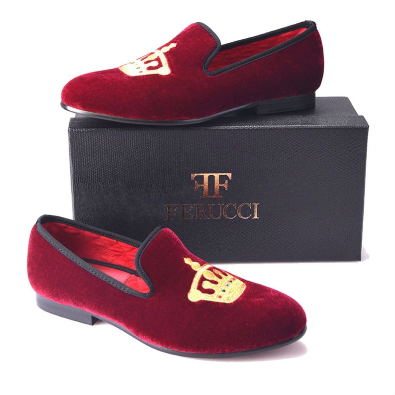May include: A pair of burgundy velvet slippers with a gold crown embroidered on the top. The slippers are on a black box with the text "TF FERRUCCI" printed on it.