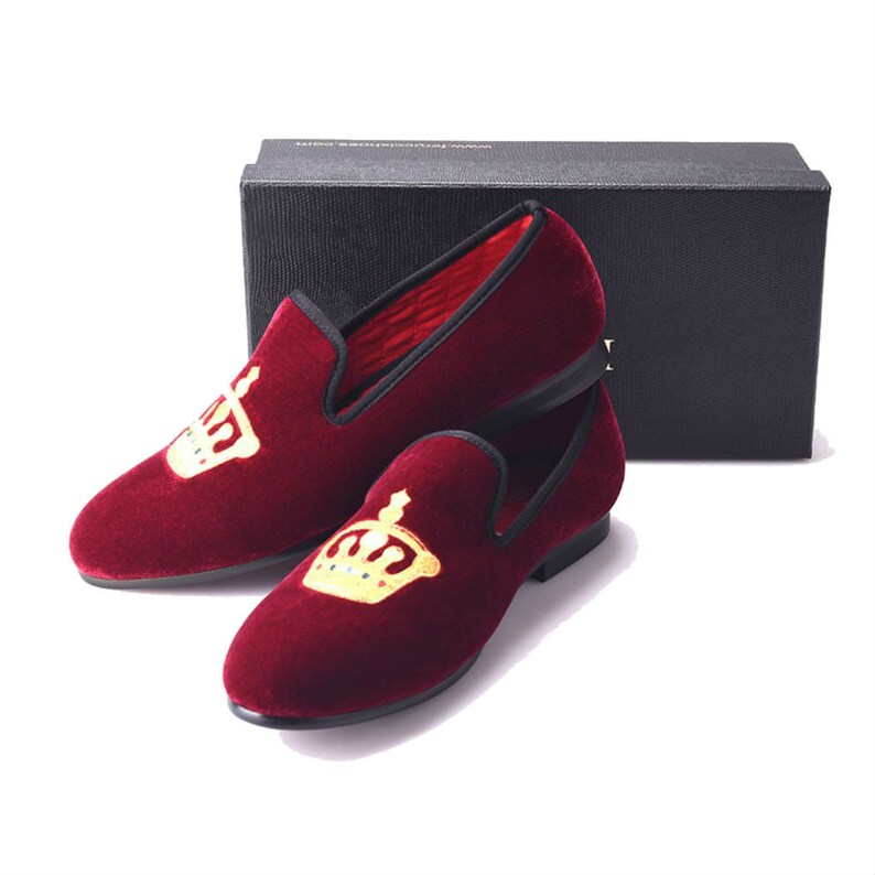 May include: A pair of burgundy velvet slippers with gold embroidered crowns. The slippers have a black trim and a black sole.