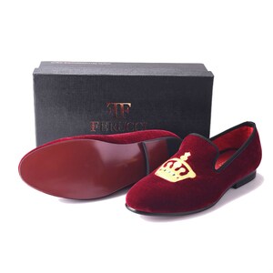 May include: A pair of burgundy velvet loafers with a gold crown embroidered on the top. The loafers are on a white background.