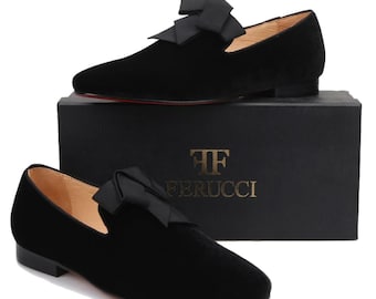 Op de afbeelding: Zwarte fluwelen loafers met een grote zwarte satijnen strik aan de bovenkant. De schoenen staan op een zwarte doos met de tekst "TF Ferrucci" in gouden letters.