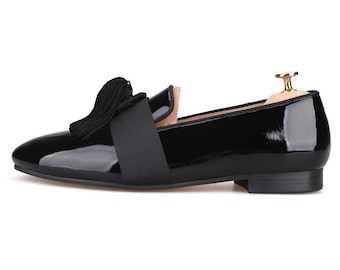 May include: Black patent leather loafer with a rounded toe and a low heel. The shoe features a black tassel and a strap across the vamp. A wooden shoe tree is inserted inside the shoe. The shoe is designed for formal or semi-formal occasions.