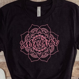 May include: Black t-shirt with a pink mandala design and the word "Survivor" in cursive. The shirt is paired with blue denim jeans and white sneakers. The items are arranged on a wooden surface.