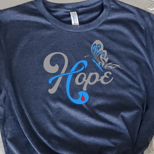 May include: A navy blue t-shirt with a gray butterfly and the word "Hope" in blue lettering. The word "Hope" has a blue teardrop underneath it.