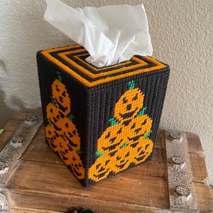 May include: A black tissue box cover with a design of stacked orange pumpkins with green stems and black eyes and mouths. The pumpkins are arranged in a pyramid shape.