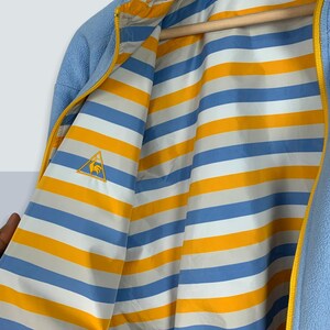 May include: A light blue fleece jacket with a yellow zipper and a striped lining. The lining is blue, white, and yellow. The jacket has a small embroidered logo of a rooster on the left side of the chest.