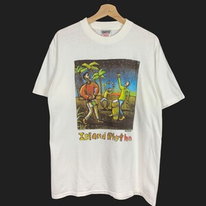 Puede incluir: Camiseta blanca con un gráfico colorido de tres figuras tocando música bajo un cielo estrellado, con palmeras. El texto "Island Rhythm" está debajo del gráfico. La camiseta está en una percha de madera.