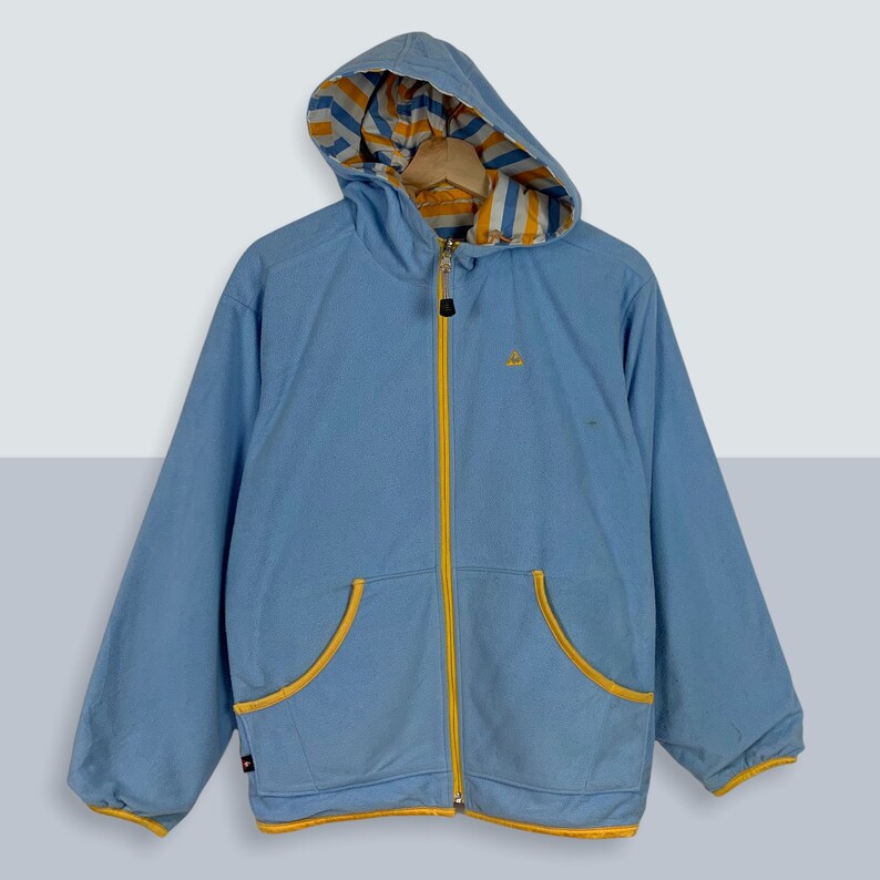 May include: A light blue hooded jacket with a yellow zipper and yellow trim. The hood is lined with a white and yellow striped fabric.
