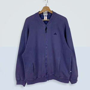 May include: A vintage purple Adidas zip-up jacket with a small Adidas logo on the chest. The jacket has a full zipper, two front pockets, and ribbed cuffs and hem. It is hanging on a wooden hanger against a white background.