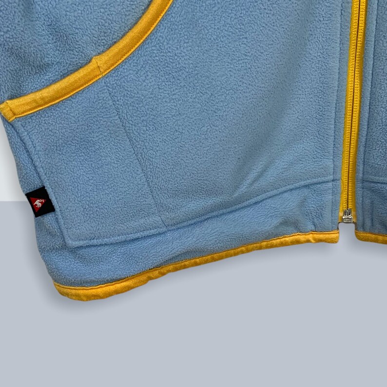 May include: A light blue fleece jacket with a yellow zipper and trim. The jacket has a small logo patch on the left side.