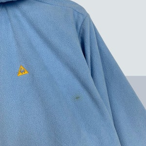 May include: A light blue fleece jacket with a yellow patch featuring a rooster logo.