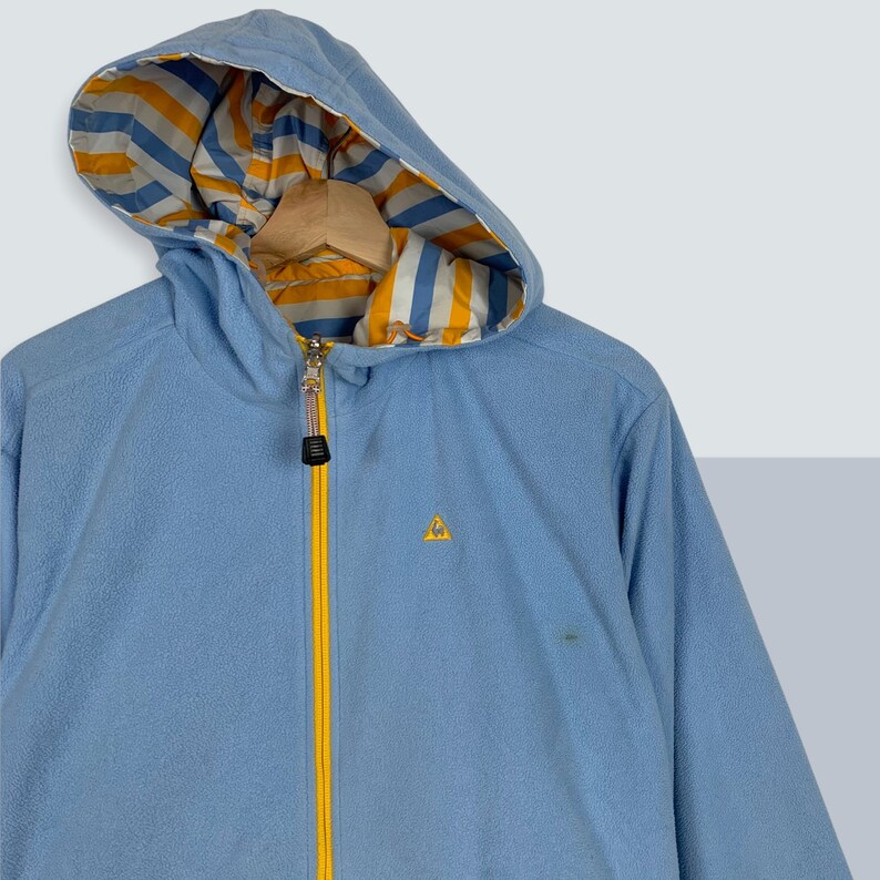 May include: A light blue fleece jacket with a yellow zipper and a yellow triangle logo. The hood is lined with a white and yellow striped fabric.