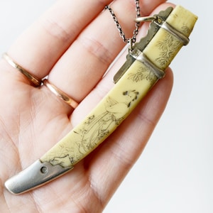 May include: A silver chain necklace with a pendant shaped like a Japanese sword. The pendant is made of ivory and features an etched design of a woman in a traditional Japanese kimono.