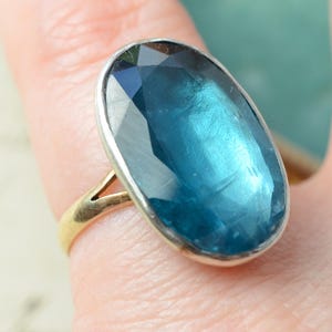 May include: A ring featuring a large, oval-shaped, faceted blue gemstone set in a silver-toned bezel. The band of the ring is made of a thin, gold-toned metal that splits into a V-shape where it connects to the base of the gemstone setting.