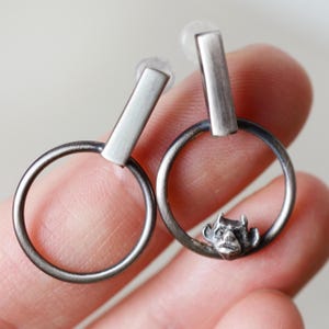 Può includere: Un paio di orecchini color argento. Ogni orecchino presenta un cerchio con un piccolo ciondolo dettagliato a forma di faccia di diavolo e una barra rettangolare sopra il cerchio. Gli orecchini sono mostrati su uno sfondo neutro.