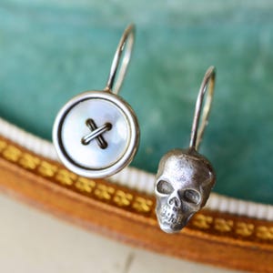 May include: A pair of silver earrings. One earring is a button design with a mother-of-pearl center and a cross-stitch detail. The other earring is a small skull. Both earrings have a curved hook.