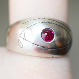 Peut inclure: Une bague en argent avec une pierre précieuse rouge sertie au centre. La bague est gravée d'un motif d'œil stylisé.