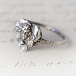 Op de afbeelding: Een zilveren ring met een bloemmotief en een gebeeldhouwd gezicht. De ring heeft een delicate band en een vintage esthetiek. De ring is waarschijnlijk gemaakt van sterling zilver. Het ontwerp van de ring is ingewikkeld.