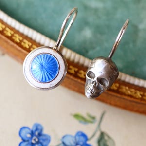 May include: A pair of silver earrings. One earring has a blue and white enamel design, the other a skull. The earrings have a hook fastening. The earrings are displayed on a floral background.