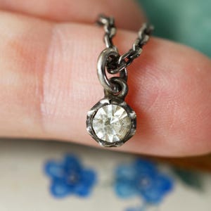Op de afbeelding: Een kleine, zilverkleurige hangerketting met een heldere, facetgeslepen edelsteen. De hanger heeft een geschulpte bezel-zetting en hangt aan een delicate ketting. De ketting wordt getoond tegen een vage achtergrond.