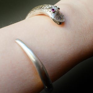 May include: A silver-toned open cuff bracelet featuring a detailed snake head terminal with a small, round, vibrant pink gemstone set as an eye. The metal surface displays a textured, scale-like pattern on the head and a smooth, polished finish.