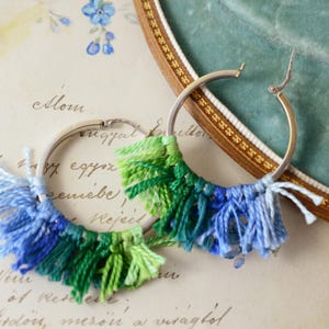 May include: A pair of silver hoop earrings with colorful fringe. The fringe is made of blue and green yarn, creating a textured and bohemian look. The earrings are displayed on a vintage background.