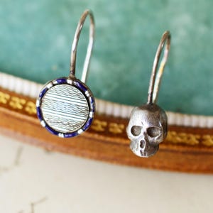 May include: A pair of silver earrings. One earring features a small skull charm, while the other has a round, patterned design with blue and white accents. The earrings have a hook closure.