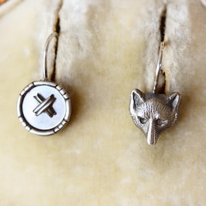 May include: A pair of silver earrings. One earring features a button design with an X across the center. The other earring is a fox head. Both earrings have a curved hook for the ear.