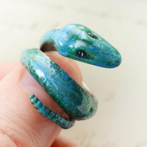 May include: A wrap-around ring shaped like a snake, featuring a glossy finish with blended shades of blue and green. The snake head is detailed with dark eyes, and the body shows a textured, scaled pattern that tapers toward the tail end.