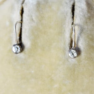 May include: A pair of silver-toned drop earrings, each featuring a single, clear, round gemstone. The earrings are displayed against a soft, light-yellow background, suggesting a jewelry box or display case. The gemstones sparkle.