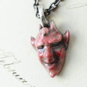 May include: A pendant shaped like a small, red, glossy devil face with pointed ears and horns, suspended from a dark, oxidized silver-toned metal chain. The facial features are sculpted with a smooth, reflective finish and deep red tones.