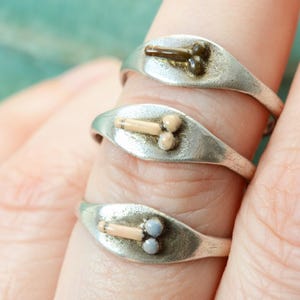 May include: Three silver rings with a phallic design. Each ring features a small, oval-shaped face with a raised, detailed design in shades of brown and beige. The rings are worn on a finger.