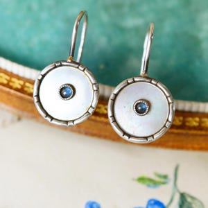 May include: A pair of silver drop earrings. Each earring has a round, white, mother-of-pearl disc with a small, blue gemstone in the centre. The earrings have a decorative, ridged border and a simple hook closure.