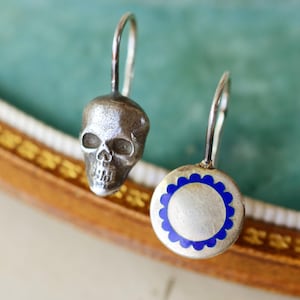 May include: A pair of silver earrings. One earring is a skull, the other is a silver disc with a blue floral border. The earrings have a hook closure.