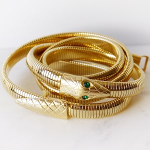 Puede incluir: Un cinturón de serpiente de metal dorado flexible con un patrón de escamas texturizado en la cabeza y la cola. La cabeza de la serpiente tiene dos pequeñas piedras redondas de color verde vibrante como ojos y el cuerpo es un eslabón continuo.