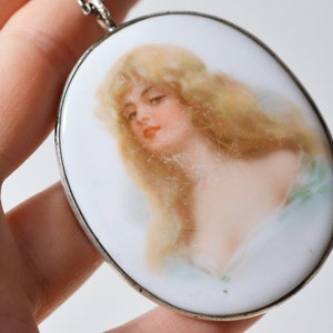 May include: A silver pendant with a white porcelain oval cameo featuring a painted portrait of a woman with long blonde hair. The woman is wearing a light blue dress and has a delicate, feminine look.