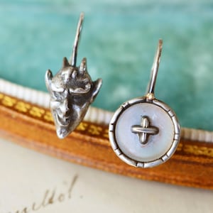 May include: A pair of silver earrings. One earring features a devil face, the other a button design with a cross. The earrings have a hook closure. The background is a soft teal colour.