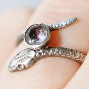 May include: A silver snake ring with a clear gemstone set in the head. The snake is coiled around the finger and has a textured surface.